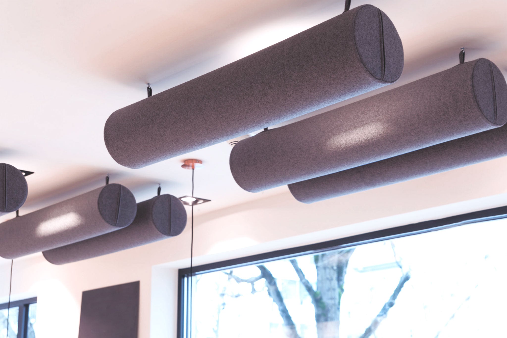 Ein Raum mit mehreren an der Decke aufgehängten Akustikzylindern und einem grossen Fenster mit viel Tageslicht.